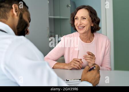Happy older mature female patient talking to indian male doctor at appointment. Stock Photo