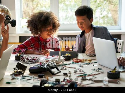 Asian school kid boy helping African girl using tablet programming robotic cars. Stock Photo
