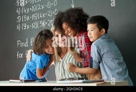 Happy diverse kids school children students hugging female teacher in classroom. Stock Photo