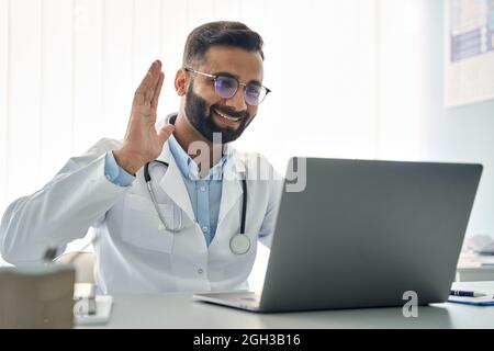 Indian male doctor waving hand having virtual online consultation on laptop. Stock Photo