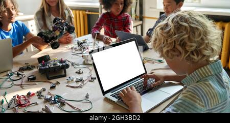 School kid boy using laptop computer with mockup screen sitting at STEM class. Stock Photo