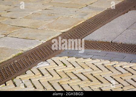 a drainage ditch covered with an iron patterned lattice on a sidewalk made of granite rough tiles stones close-up of a stormwater drainage system from Stock Photo