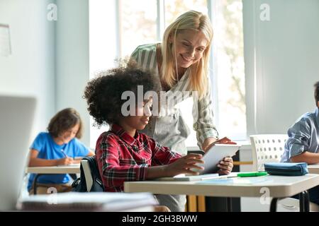 Female teacher helping African school girl using tablet at class in classroom. Stock Photo