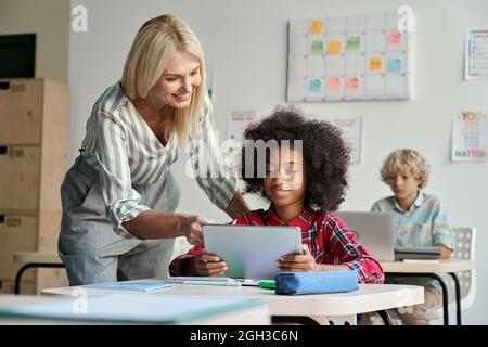 Female teacher helping African school student girl using tablet in classroom. Stock Photo
