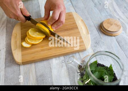 A woman cuts a lemon on a wooden kitchen board with a sharp large knife. Healthy food concept. The process of making tea with mint and lemon. Stock Photo