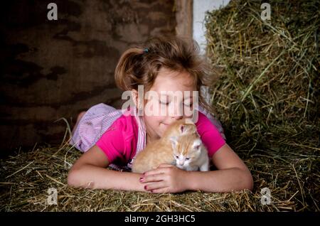 young overalls, in pink cuddles with kittens in the barn on bales of straw Stock Photo