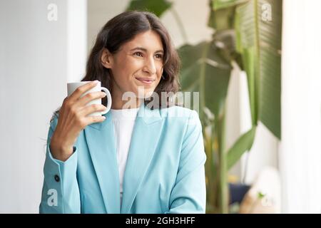 Young smiling Latina business woman, satisfied with a job well done, relaxing with her morning coffee or tea, looking out the window. Stock Photo