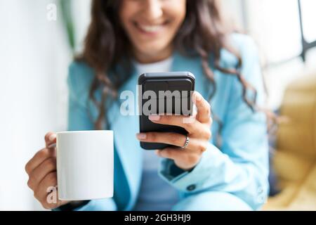 Close-up of hands of unrecognizable business woman drinking coffee sitting on a sofa while using a smart phone. Smiling working woman sends text Stock Photo