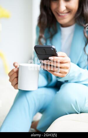 Close-up of unrecognizable business woman's hands holding a cup of coffee sitting on a sofa while using a smart phone. Vertical photo. Stock Photo
