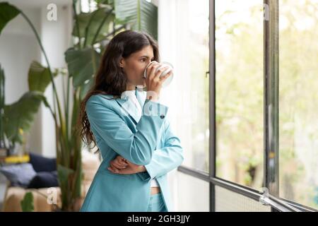 Young businesswoman enjoying her morning coffee or tea, looking out the window. Beautiful latina woman drinking hot drinks in the office. Stock Photo