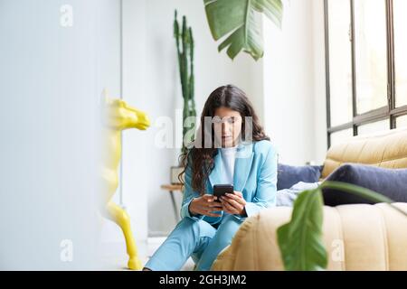 Beautiful Latina businesswoman drinking coffee in a nice modern apartment using a smartphone enjoying relaxed browsing messages planning to send text Stock Photo