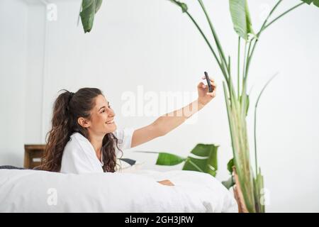 Pretty young latina woman takes a selfie lying on the bed. Influencer sharing on social networks and online connected people. Stock Photo