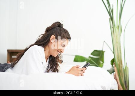 Young smiling Hispanic woman uses smart phone lying on bed. People using social networks and chatting connected by technology. Stock Photo