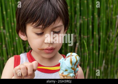 Brunette little boy eating ice cream cone with a spoon wearing sleeveless shirt in summertime. Green plants on background. Horizontal Stock Photo