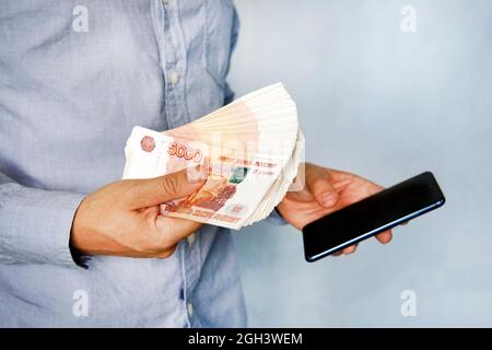 young man holds the money in his hand and presses the screen of the smartphone. Earns money through the mobile app. Business via smartphone. Stock Photo