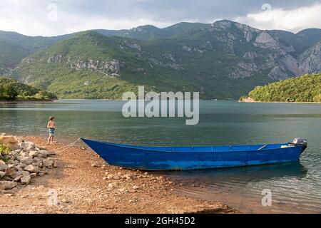 Boy fishing on a Vau i Dejes Reservoir near traditional blue boat not far away from the city of Shkodra in Albania Stock Photo