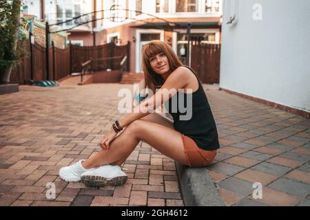 Pretty smiling young woman with tattooes is sitting on the sidewalk. Side view. The concept of relax and psyhology. Stock Photo