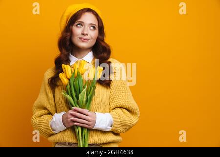 Smiling young woman with long red hair in sweater and beret standing holding tulips bouquet over yellow wall background Stock Photo