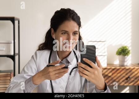 Happy focused female doctor in earphones using mobile phone Stock Photo