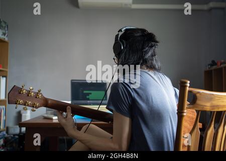 A man with long hair wearing headphones and playing acoustic guitar in order to recording into a computer in a domestic room Stock Photo