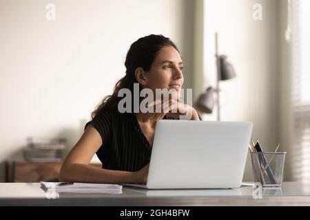 Smiling thoughtful female employee working at laptop from home Stock Photo