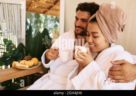 Beautiful multi-ethnic couple wearing white bathrobes drinking relaxing tea at spa. Romantic date at spa resort after relax and massage Stock Photo