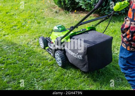 An unidentifiable adult female cutting the grass and mowing a green lawn with a Greenworks 40v battery operated cordless lawnmower. Stock Photo