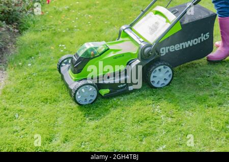 An unidentifiable adult female cutting the grass and mowing a green lawn with a Greenworks 40v battery operated cordless lawnmower. Stock Photo