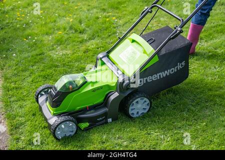 An unidentifiable adult female cutting the grass and mowing a green lawn with a Greenworks 40v battery operated cordless lawnmower. Stock Photo