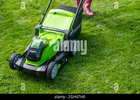 An unidentifiable adult female cutting the grass and mowing a green lawn with a Greenworks 40v battery operated cordless lawnmower. Stock Photo