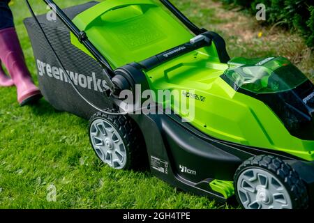 An unidentifiable adult female cutting the grass and mowing a green lawn with a Greenworks 40v battery operated cordless lawnmower. Stock Photo