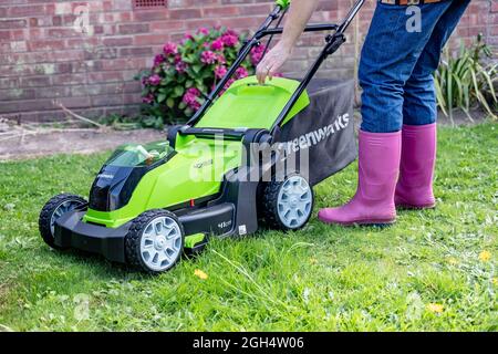 An unidentifiable adult female cutting the grass and mowing a green lawn with a Greenworks 40v battery operated cordless lawnmower. Stock Photo