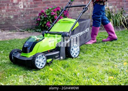 An unidentifiable adult female cutting the grass and mowing a green lawn with a Greenworks 40v battery operated cordless lawnmower. Stock Photo