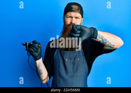 Redhead man with long beard tattoo artist wearing professional uniform and gloves looking unhappy and angry showing rejection and negative with thumbs Stock Photo