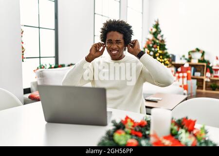 Young african american man using laptop sitting on the table by christmas tree covering ears with fingers with annoyed expression for the noise of lou Stock Photo