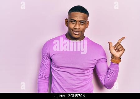 Young black man wearing casual pink sweater with a big smile on face, pointing with hand finger to the side looking at the camera. Stock Photo