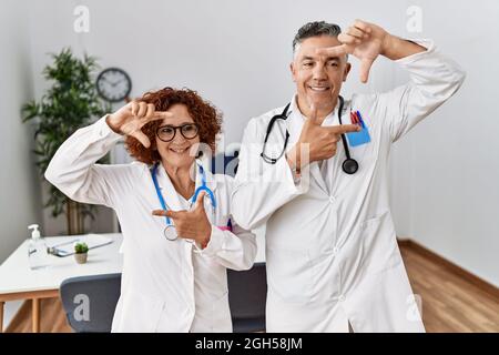 Two middle age doctors at medical clinic smiling making frame with hands and fingers with happy face. creativity and photography concept. Stock Photo