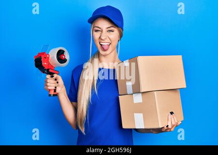 Young caucasian woman holding packages and packing tape sticking tongue out happy with funny expression. Stock Photo