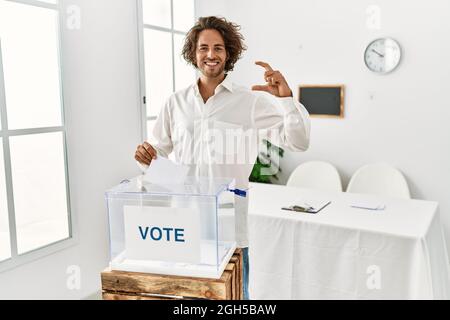 Young hispanic man voting putting envelop in ballot box smiling and confident gesturing with hand doing small size sign with fingers looking and the c Stock Photo