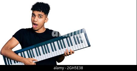 Young african american man holding piano keyboard in shock face, looking skeptical and sarcastic, surprised with open mouth Stock Photo