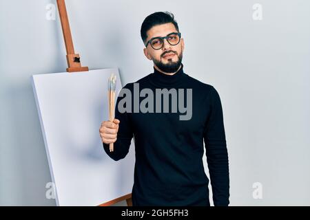 Handsome man with beard holding brushes close to easel stand looking sleepy and tired, exhausted for fatigue and hangover, lazy eyes in the morning. Stock Photo