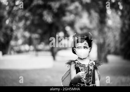 A grayscale shot of a Southeast Asian girl wearing her costume and also a facemask on Halloween, outdoors Stock Photo