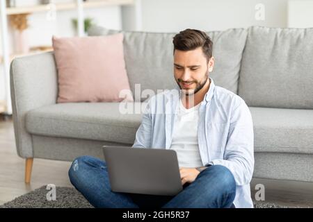Smiling millennial man working from home, sitting on floor and using laptop, free space Stock Photo