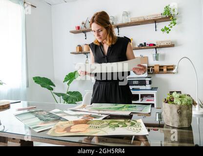 Creative female designer examining various paintings placed on table and working on project Stock Photo