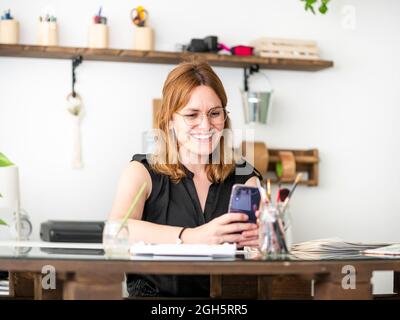 Cheerful female designer using smartphone while sitting in table and working in creative workspace Stock Photo