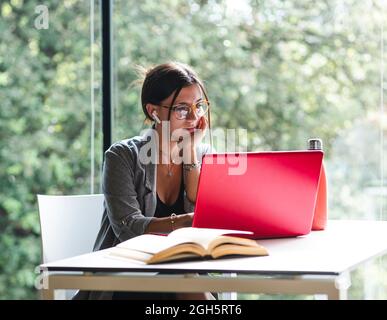 Side view of female student typing on netbook while sitting at table with book and doing homework in university library Stock Photo