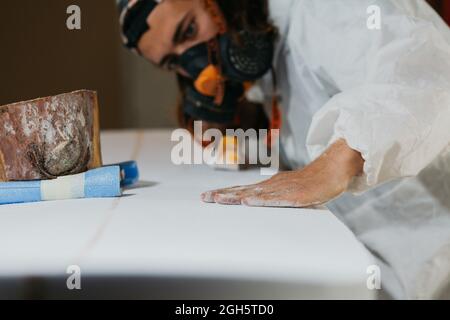 Male shaper in respirator and safety costume smoothing surface of surfboard during shaping process in workshop Stock Photo