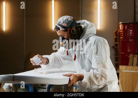Side view of male shaper in respirator and protective costume using plane on surfboard surface in workshop Stock Photo