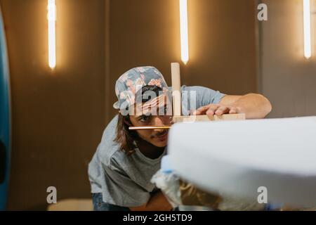 Concentrated male hipster using scribe tool with pencil while marking surfboard before shaping in workshop Stock Photo