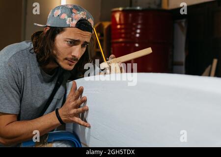 Concentrated male hipster using scribe tool with pencil while marking surfboard before shaping in workshop Stock Photo